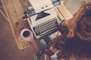 Above view of curly woman thinking and taking coffee with old typewriter on the table is the featured image for the blog article by kelley way titles