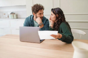 A couple in their mid-thirties looking worriedly at each other while in front of a laptop with a document in the womens hand for featured image for blog article by Kelley Way titled The Top 5 Estate Planning Mistakes People Make